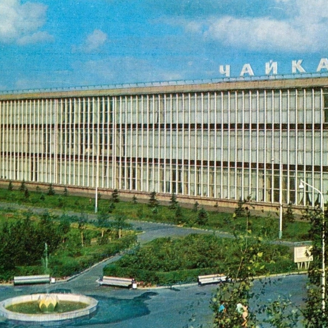 Советское здание с надписью Чайка на крыше, окруженное зелеными насаждениями и дорожками, на фоне голубого неба.
